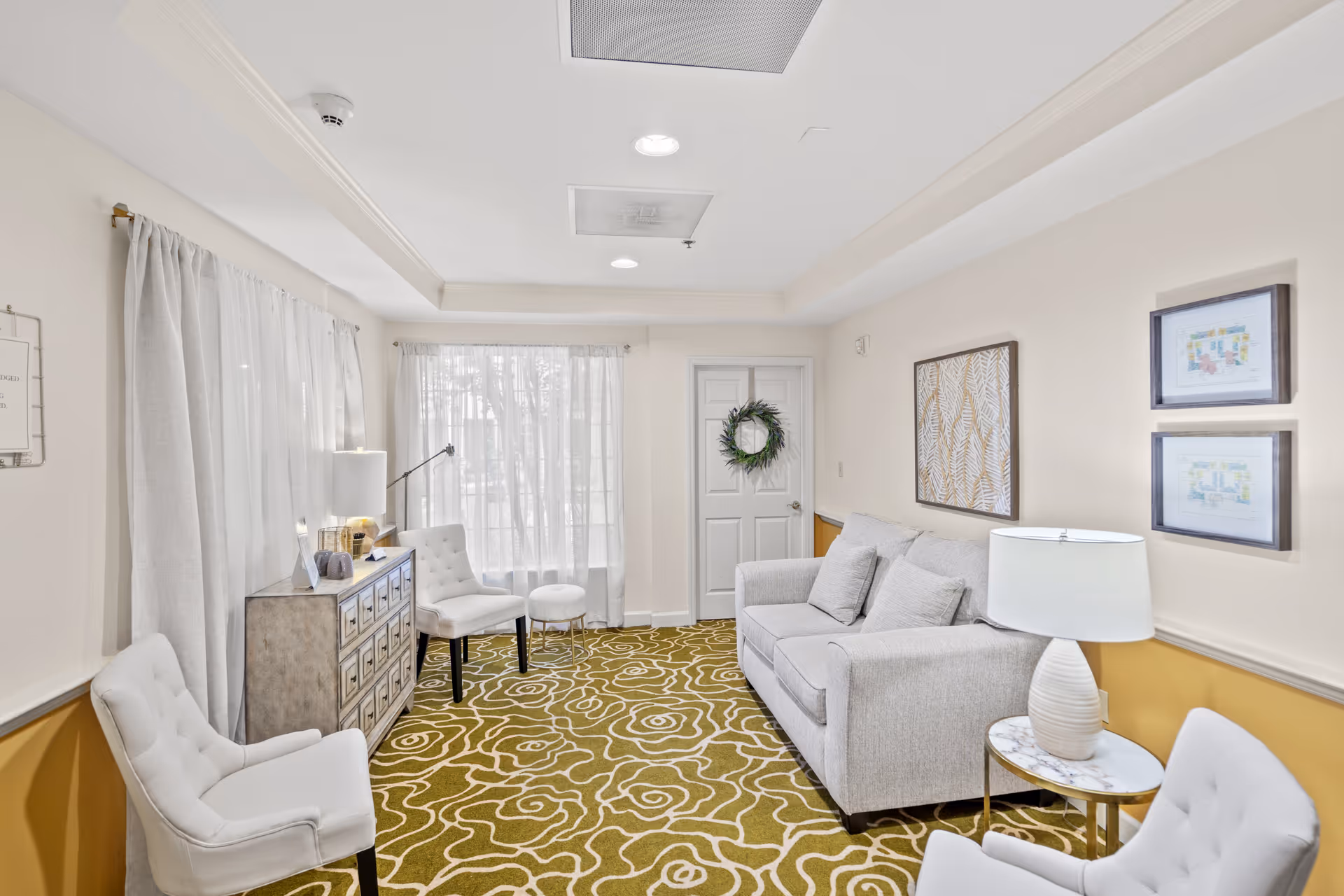 A bright and cozy living room with a patterned green carpet, a gray sofa with cushions, two white upholstered chairs, a small round side table with a white lamp, a wooden chest of drawers, and large windows with sheer white curtains letting in natural light. The walls are decorated with framed artwork and a wreath on a white door.