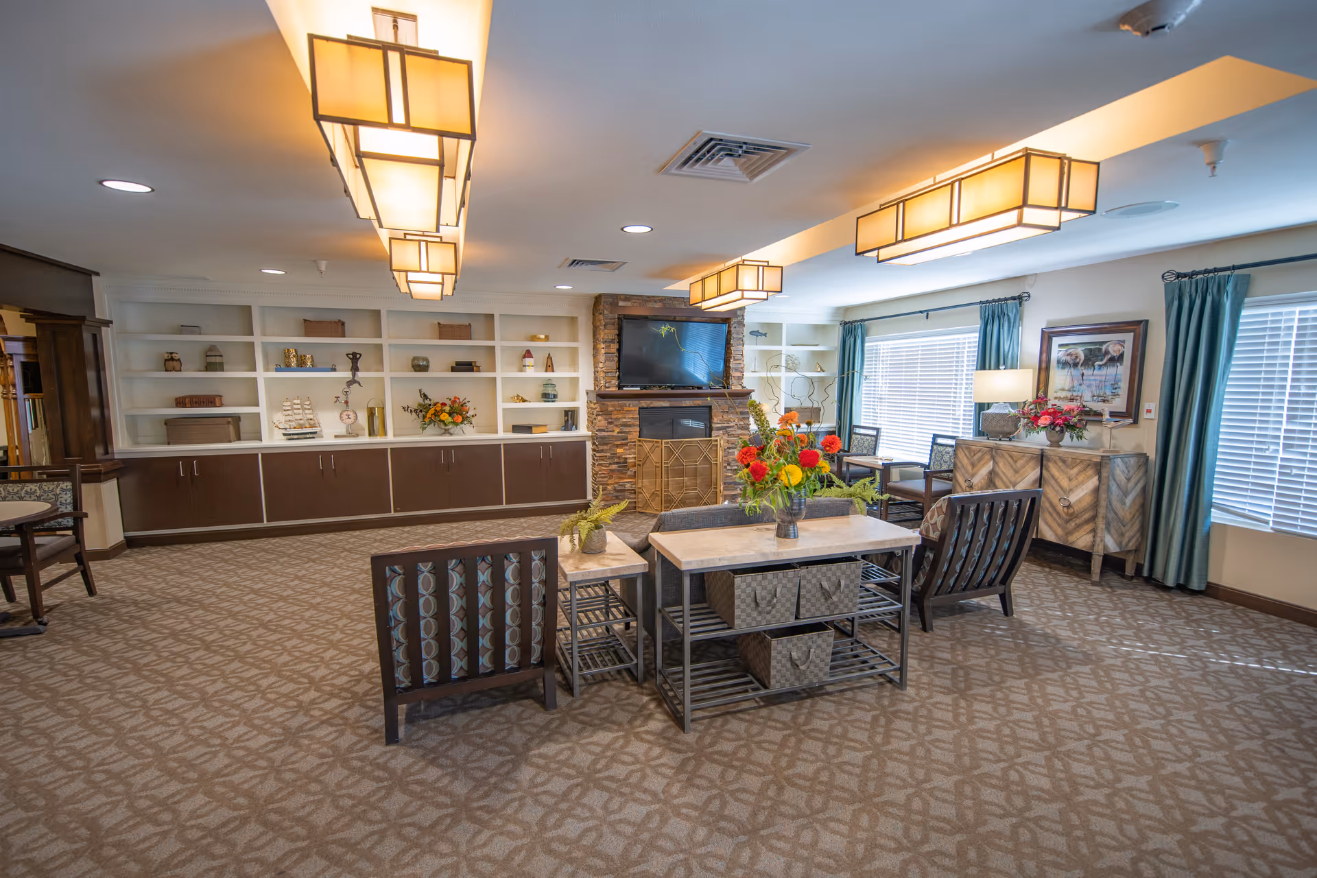 A spacious and well-lit senior living community lounge featuring patterned carpet, modern rectangular ceiling lights, a stone fireplace with a mounted TV above, and built-in shelves with decorative items. The room has multiple seating areas with cushioned chairs and tables, large windows with teal curtains, and floral arrangements on tables and a sideboard.