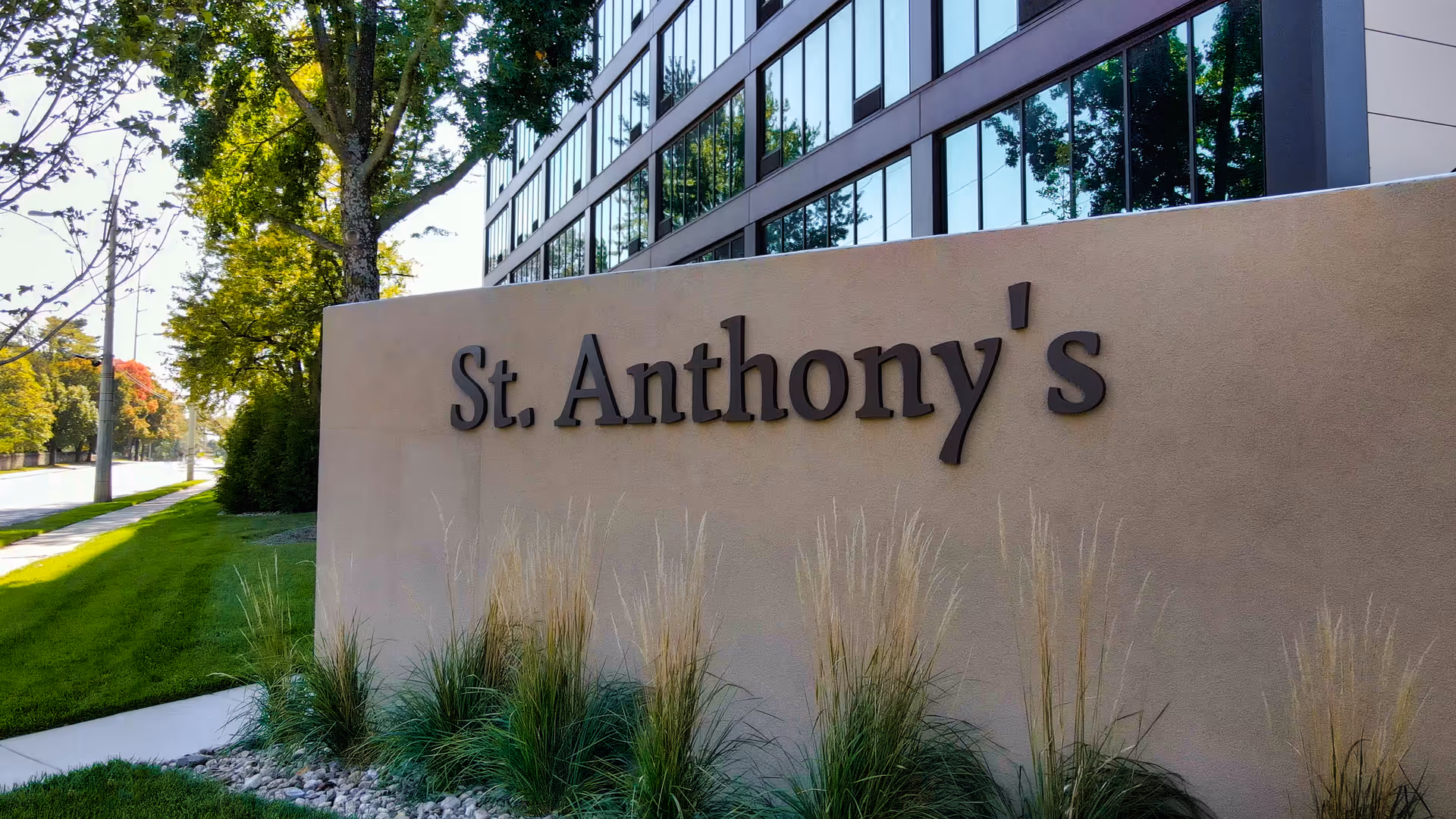 Exterior view of a building with a beige wall sign that reads 'St. Anthony's' surrounded by ornamental grasses and trees along a sidewalk.