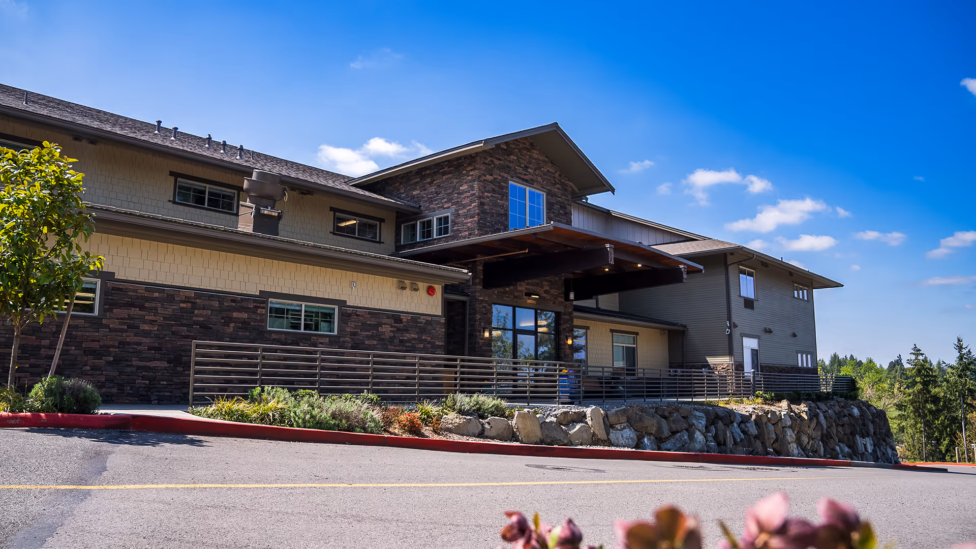 Front exterior of a two-story memory care building with stone and siding facade, entrance canopy, landscaped rock wall and a clear blue sky.