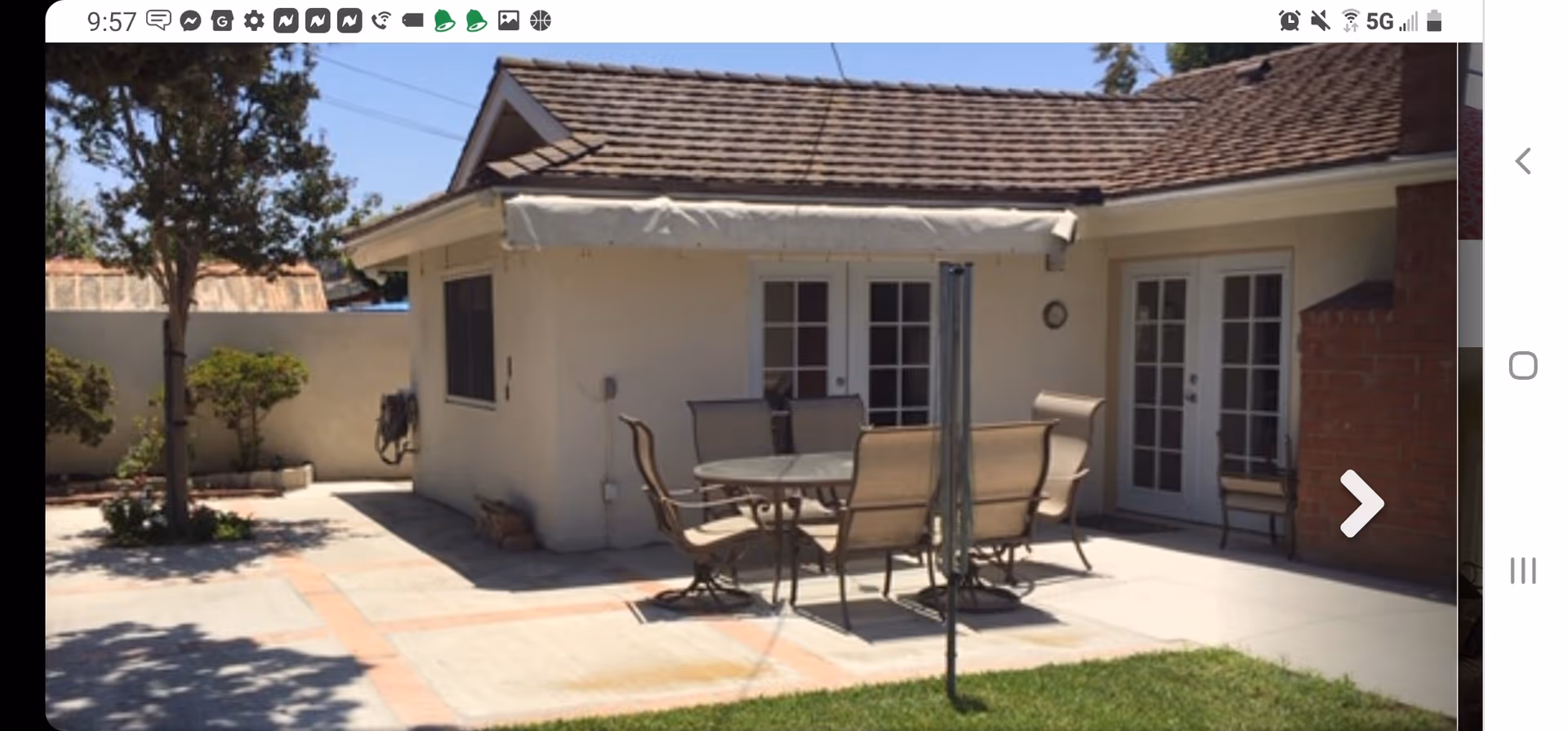 Outdoor patio area with a round glass table and six beige chairs on a concrete surface. The patio is adjacent to a light-colored building with two sets of French doors and a small awning above one door. There is a tree and some shrubs along the perimeter wall, and a grassy lawn in the foreground.