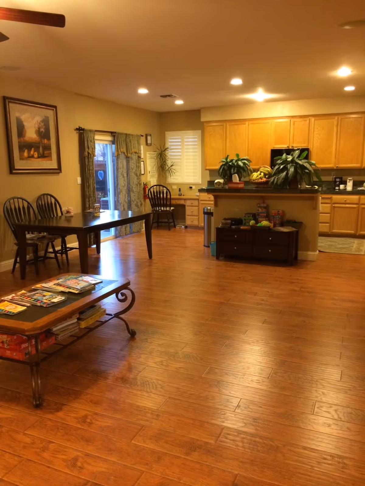 Interior view of a senior living facility showing a spacious room with wooden flooring. There is a wooden dining table with chairs near a sliding glass door with curtains. A coffee table with magazines and board games is in the foreground. The background features a kitchen area with wooden cabinets, a countertop with plants and fruit, and a small desk with a chair. The room is well-lit with recessed ceiling lights.