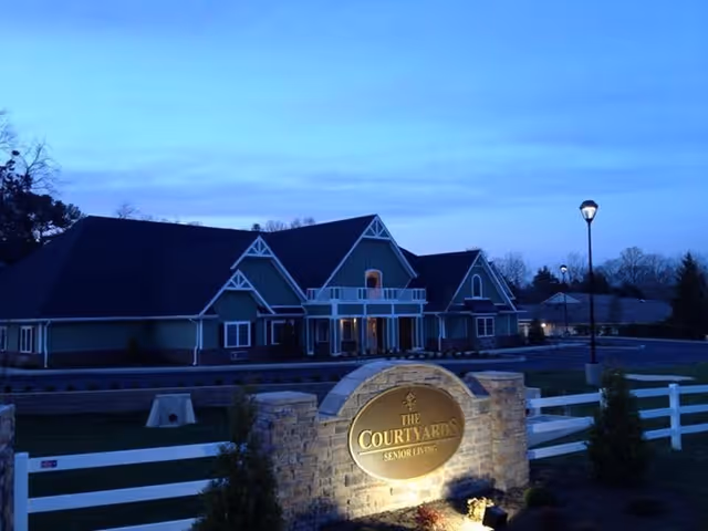 Exterior view of The Courtyards Senior Living facility at dusk, showing a large building with multiple peaked roofs and a well-lit stone sign in the foreground. The area is surrounded by a white fence and some landscaping, with a streetlamp illuminating the parking area.