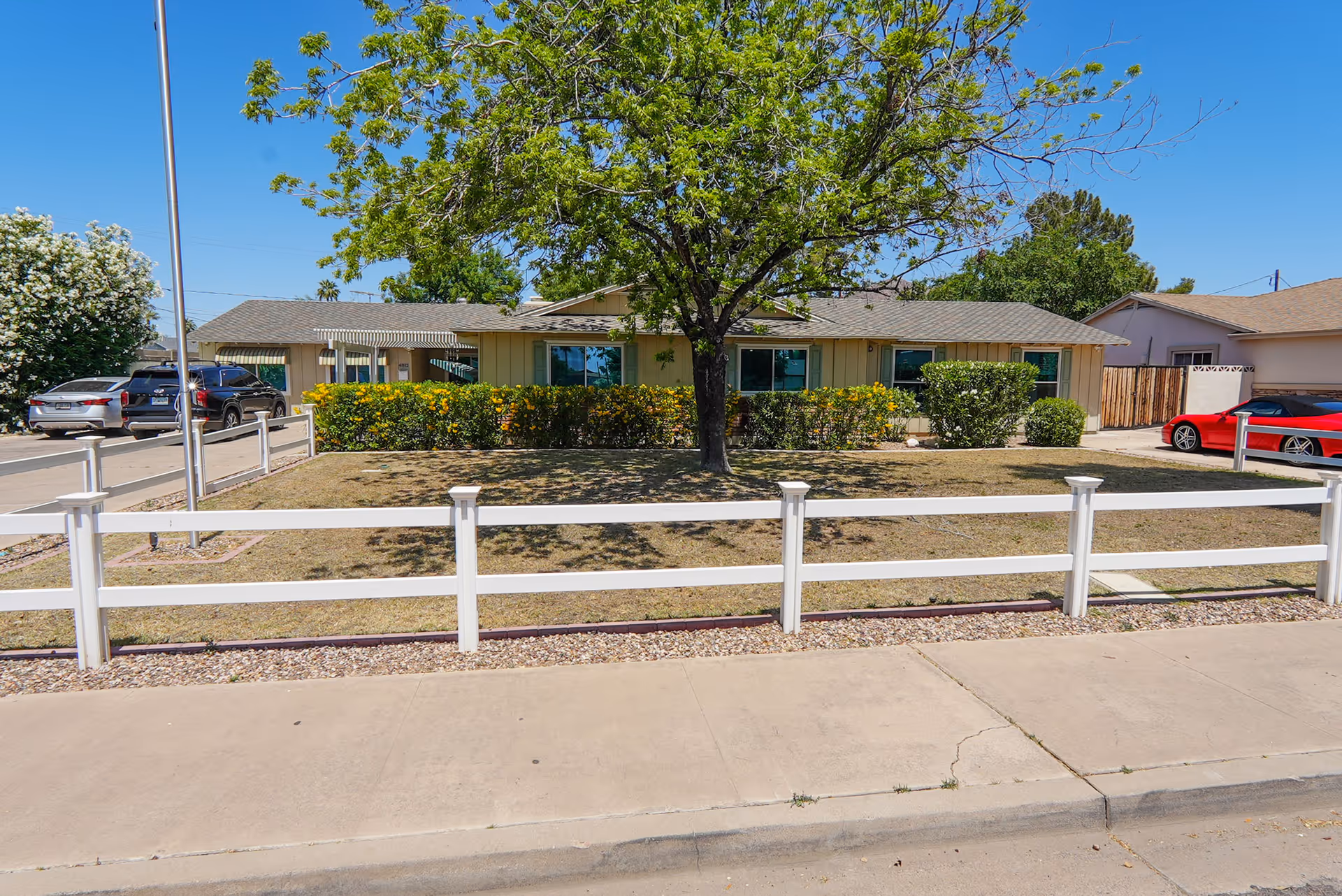 Single-story residential building with a beige exterior, surrounded by a white fence and a front yard with a tree and bushes. Several cars are parked on the driveway and street under a clear blue sky.