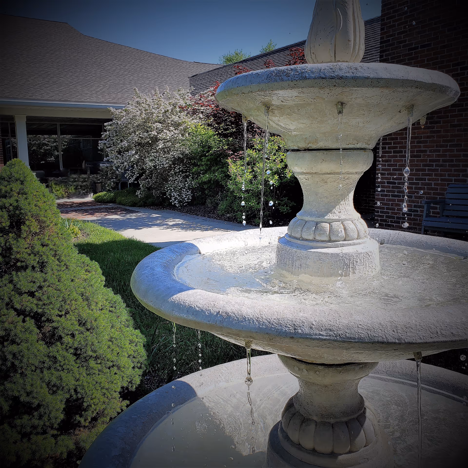 A close-up view of a two-tiered stone water fountain with water flowing from the top tier to the bottom tier. In the background, there is a brick building, a paved walkway, green bushes, and flowering shrubs under a clear blue sky.