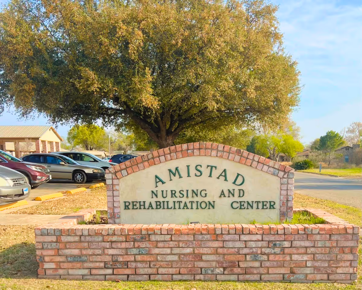 Brick sign for Amistad Nursing and Rehabilitation Center with a large tree and parked cars in the background under a clear sky.