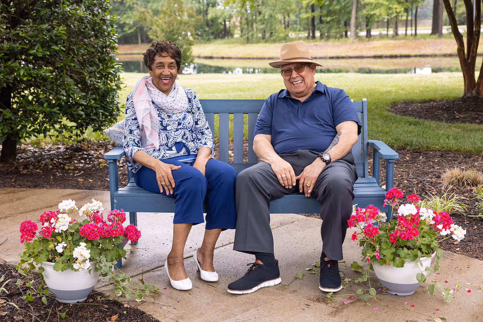 An elderly man and woman sitting on a blue bench outdoors, smiling and enjoying a garden setting with green grass, trees, and a pond in the background. There are two white pots with pink and white flowers placed on either side of the bench.