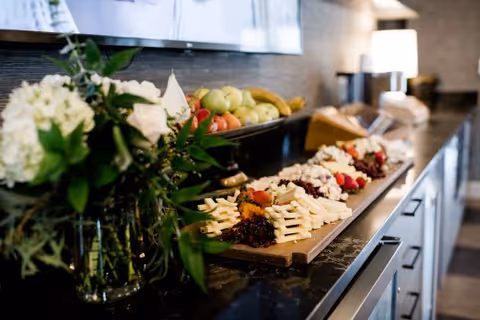 A buffet counter topped with a charcuterie and cheese board, fresh fruit, and a vase of flowers in a communal dining area.