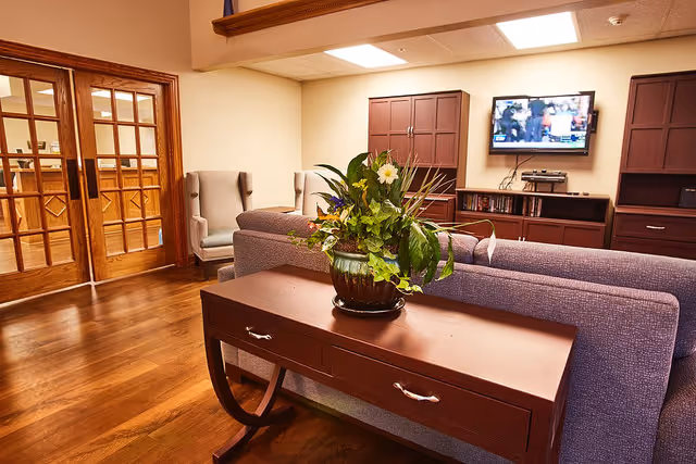A cozy living room area with a purple sofa, a wooden console table with a potted plant, two armchairs, wooden cabinets, and a wall-mounted TV showing a sports game. The room has wooden flooring and beige walls with a ceiling light.