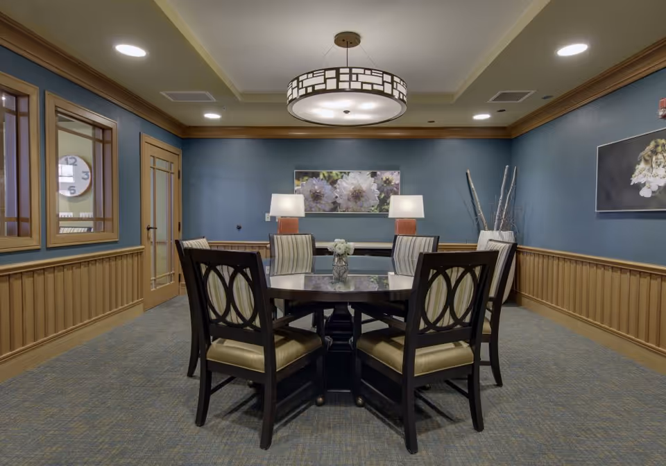 Small dining/meeting room with a round table surrounded by chairs, two table lamps, and floral artwork on blue walls.
