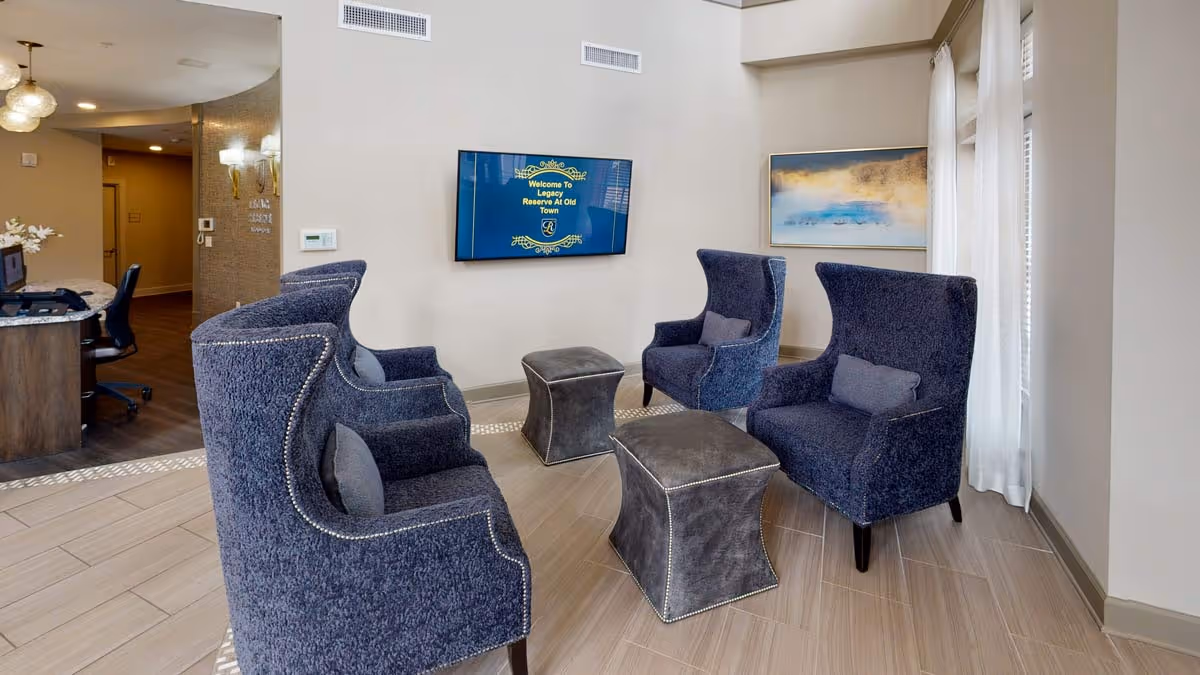 A bright lobby lounge with four navy wingback chairs and two ottomans arranged around a wall-mounted TV, with a reception desk visible to the left.