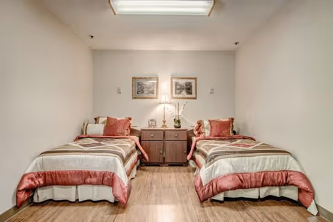 A simple bedroom with two twin beds separated by a wooden nightstand. Each bed has a beige and burgundy comforter with matching pillows. A lamp and decorative items are on the nightstand, and two framed pictures hang on the wall above it. The room has light-colored walls and wooden flooring.