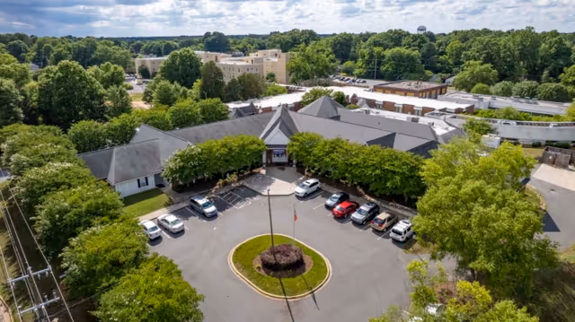 Aerial view of Rock Hill Post Acute Care Center showing a large building with a circular driveway and several parked cars. The building is surrounded by many trees and greenery, with a clear sky above.