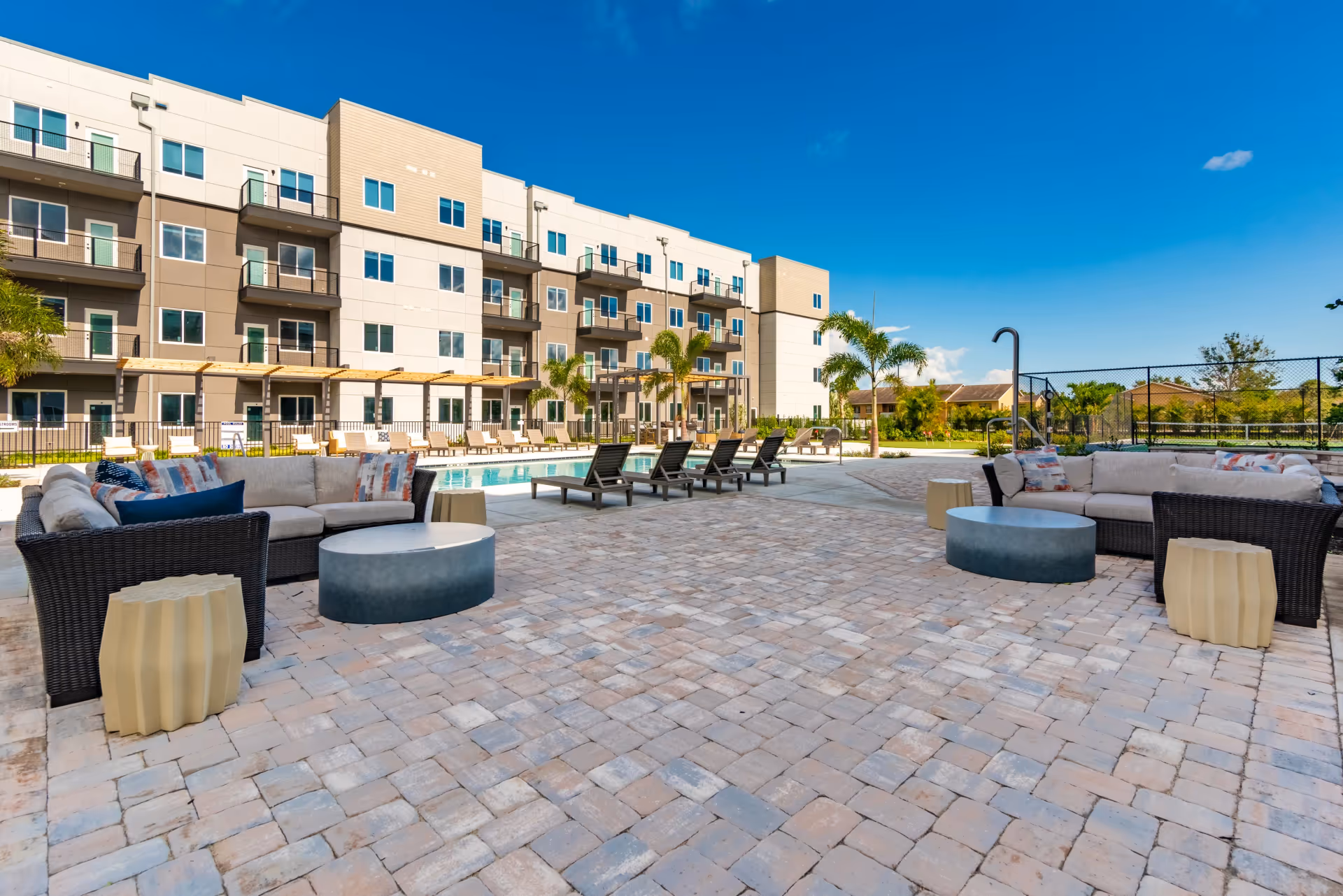 Outdoor patio area with cushioned wicker sofas, round tables, and lounge chairs surrounding a swimming pool in front of a multi-story residential building under a clear blue sky.