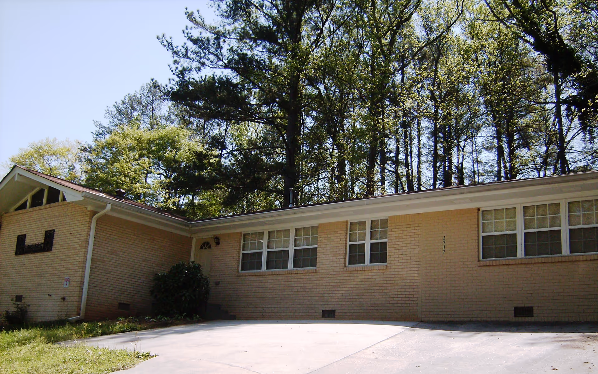 A single-story brick building with large windows sits along a paved driveway, surrounded by trees.