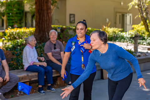An elderly woman in a blue sweater is playing a game outdoors, throwing a bean bag or similar object. A caregiver in a blue uniform stands nearby watching and smiling. In the background, three other elderly individuals are seated on a bench, observing the activity. The setting is a garden or courtyard area with trees and shrubs.