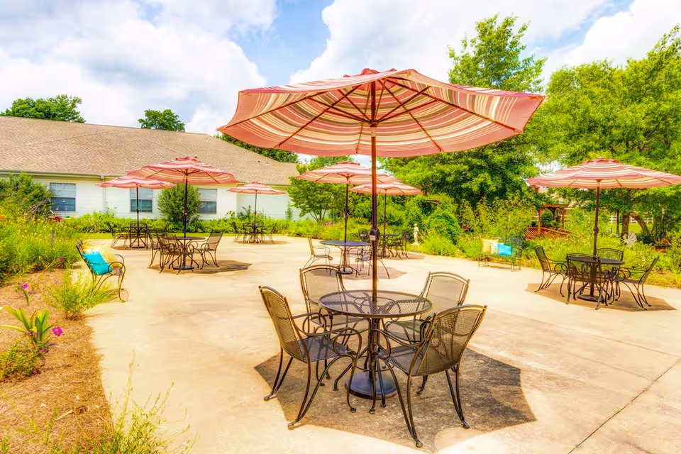 Outdoor patio area with multiple round metal tables and chairs, each shaded by large striped umbrellas. The patio is surrounded by greenery, trees, and a building with a sloped roof in the background under a partly cloudy sky.