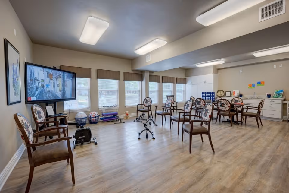A spacious activity room with wooden flooring and beige walls, featuring several floral-patterned chairs arranged around the room. There is a large flat-screen TV mounted on the wall showing an image of a street scene. The room has multiple windows with blinds, exercise equipment, and a table with chairs near the back. Cabinets and art supplies are visible along the far wall.