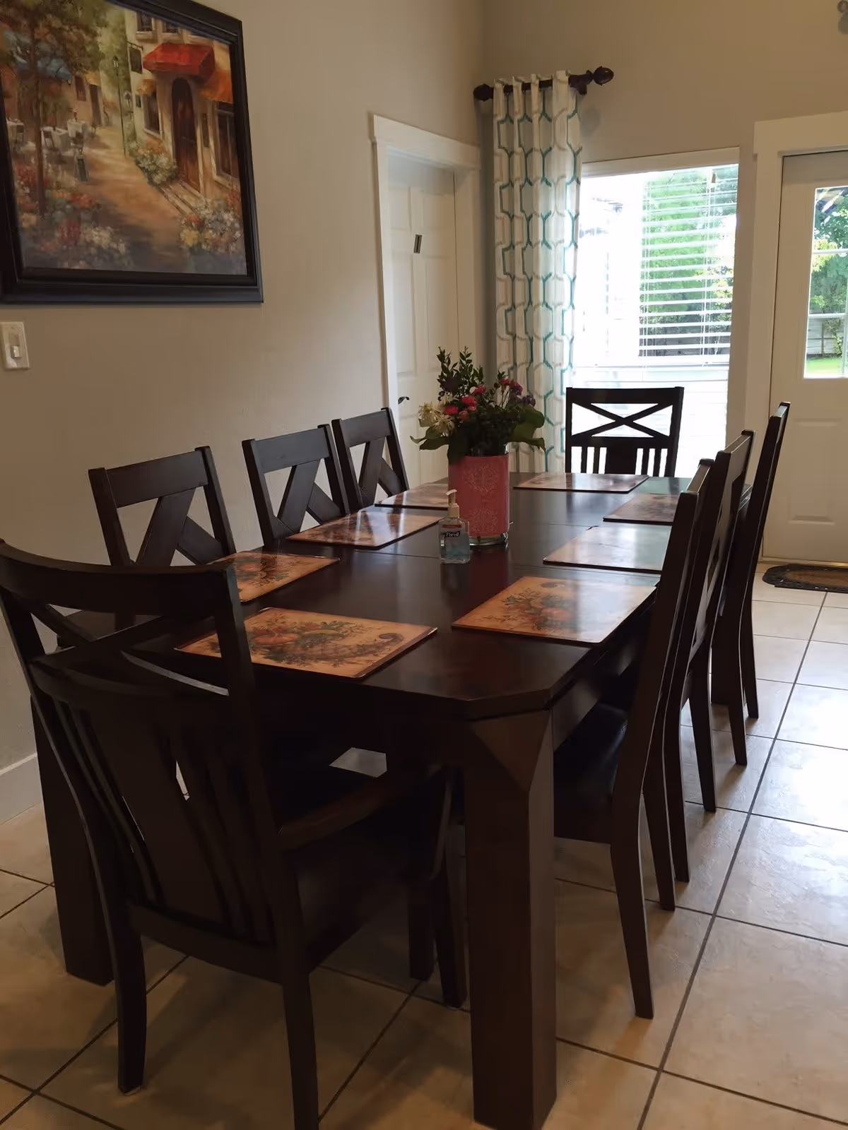 A dining room with a dark wooden rectangular table set with eight matching chairs. Each place at the table has a floral placemat. A pink vase with a bouquet of flowers and a bottle of hand sanitizer are placed in the center of the table. The room has tiled flooring, a painting on the wall, a door, and a window with patterned curtains letting in natural light.