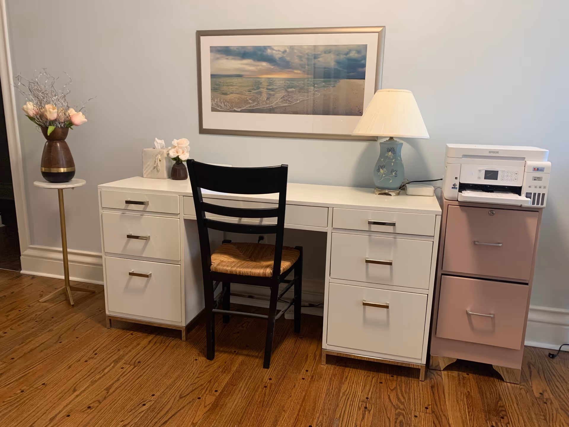 A white desk with drawers and a black chair sits against a wall beneath a framed beach print, flanked by a lamp, printer on a pink filing cabinet, and a vase of flowers on a hardwood floor.
