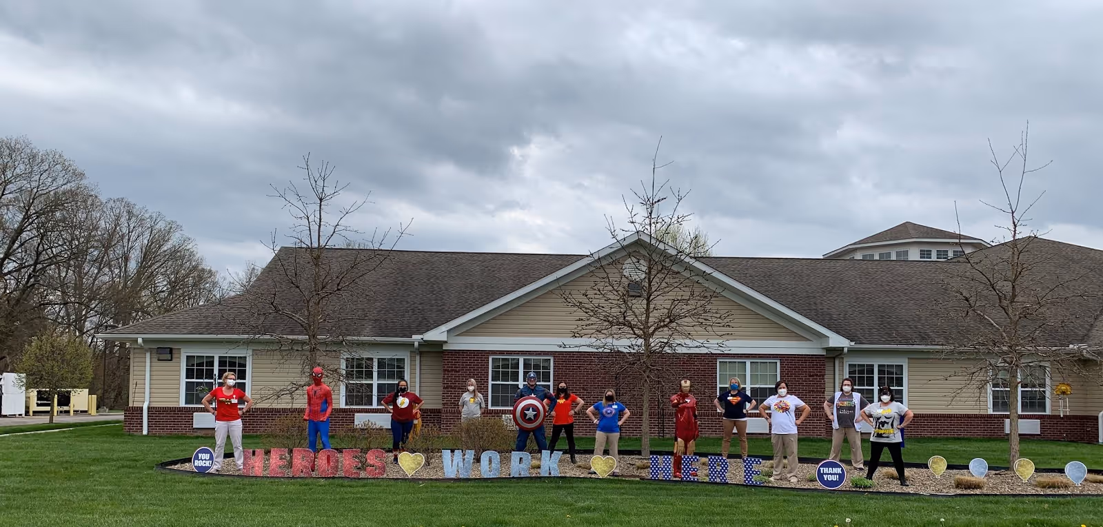 Staff wearing superhero costumes and masks stand spaced along the lawn in front of a one-story senior living building with a decorative 'Heroes Work Here' display.