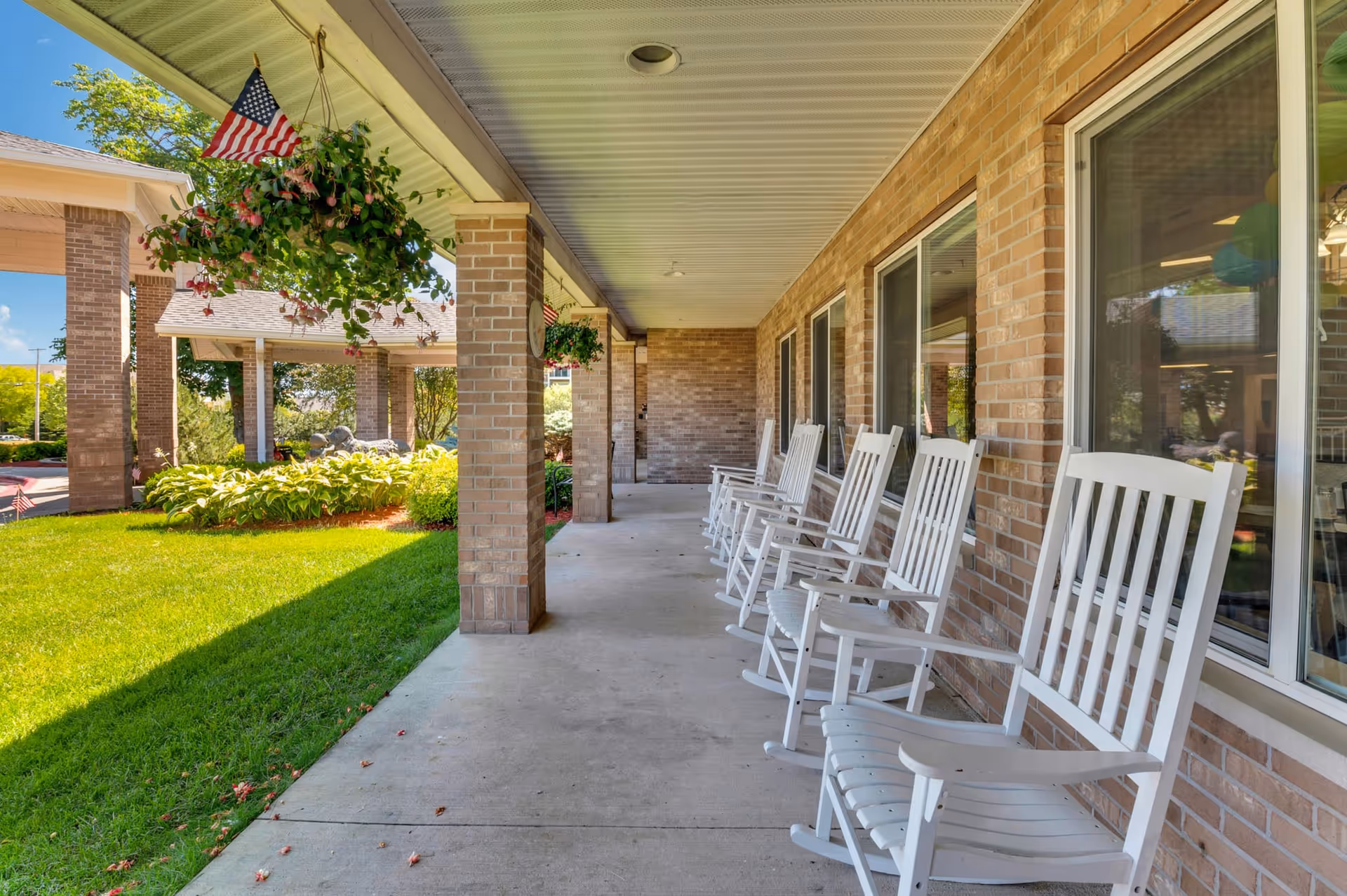 Covered front porch with a row of white rocking chairs along a brick wall, hanging plants, and a manicured lawn.