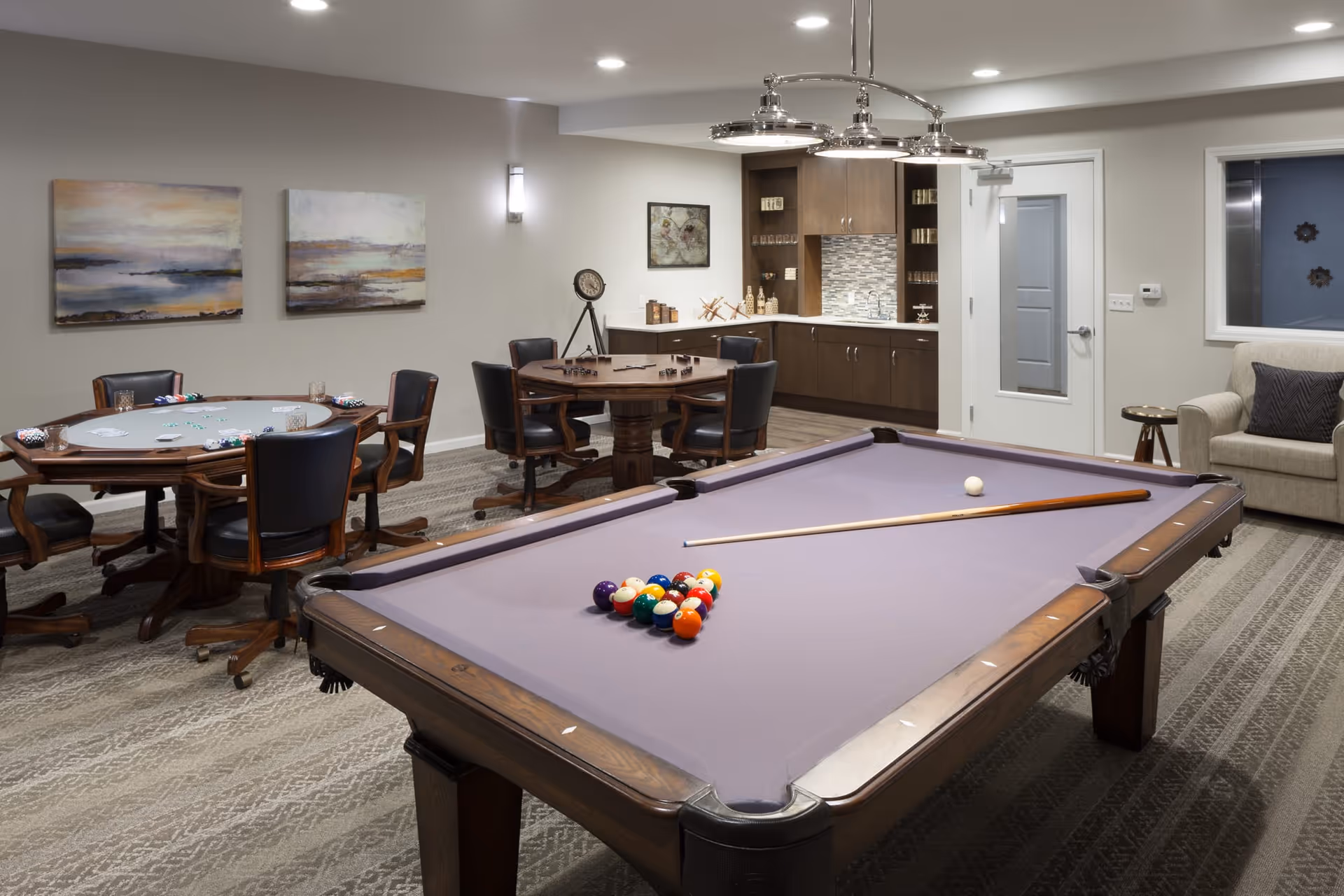 A recreational room featuring a pool table with a cue stick and balls arranged for a game. In the background, there are two round tables with chairs, one set up for card games and the other for board games. The room has modern lighting, abstract wall art, a beige armchair, and a small kitchenette area with cabinets and a countertop.