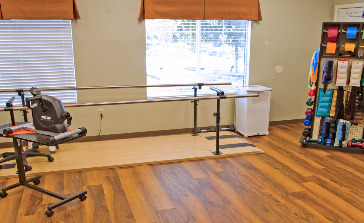 A rehabilitation or physical therapy room with parallel bars for walking exercises, a stationary exercise bike on a stand, a small white refrigerator, and a rack holding various colorful exercise and therapy equipment. The room has wood-patterned flooring and two windows with blinds and brown valances.