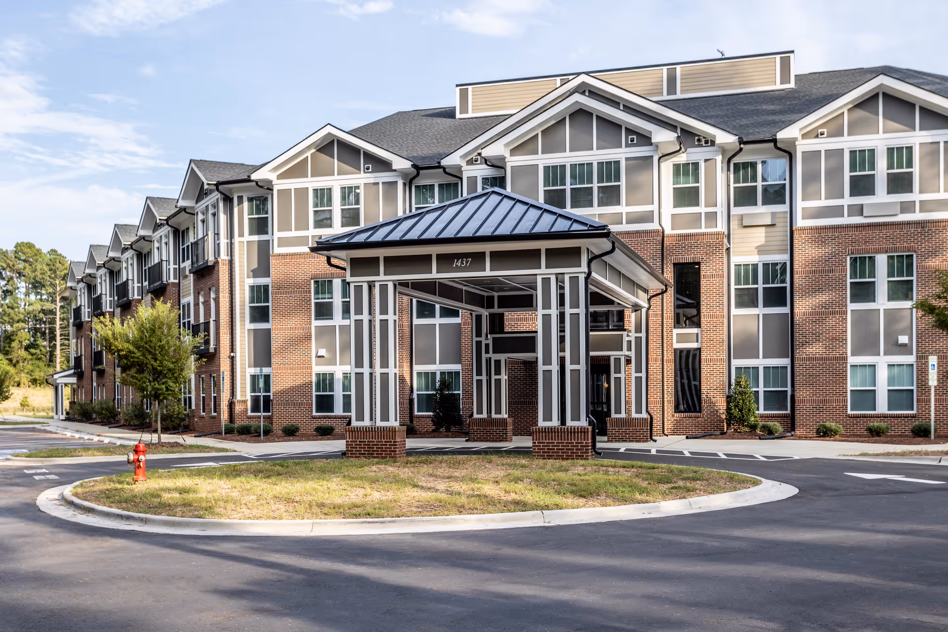 Exterior view of a multi-story senior living facility building with a covered entrance and a circular driveway. The building features a combination of brick and light-colored siding with multiple windows. There is a fire hydrant on the grass island in front of the entrance, and trees and shrubs are planted around the building.