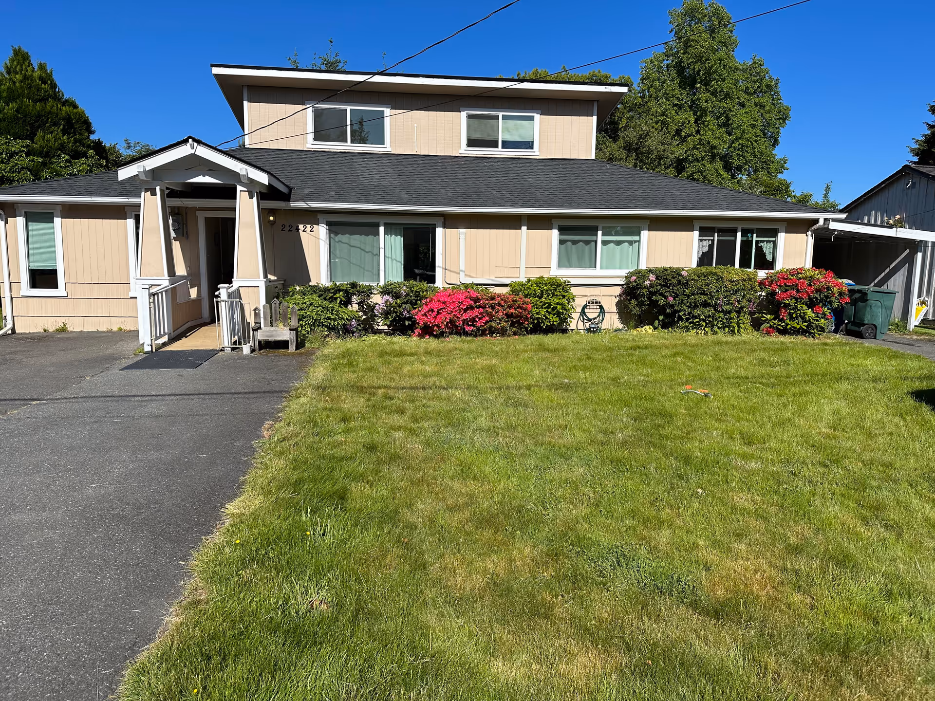 Front exterior view of a beige two-story house with a black roof, a small covered porch entrance, green bushes, and colorful flowers in front. There is a well-maintained lawn and a paved driveway on the left side under a clear blue sky.