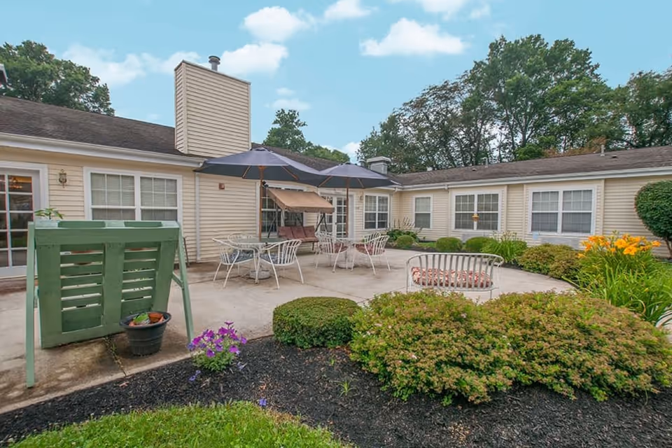 Outdoor patio area at The Courtyard at Newark featuring a concrete seating area with metal chairs and tables under two large umbrellas, surrounded by bushes, flowers, and a beige building with multiple windows in the background under a partly cloudy sky.