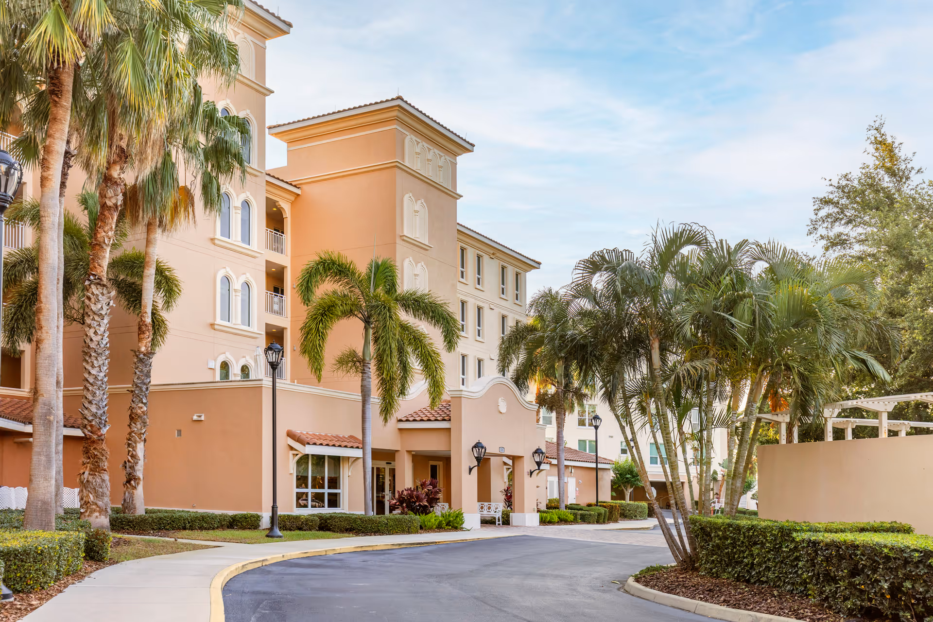 Exterior view of a multi-story senior living facility building with a curved driveway, palm trees, and well-maintained landscaping under a partly cloudy sky.