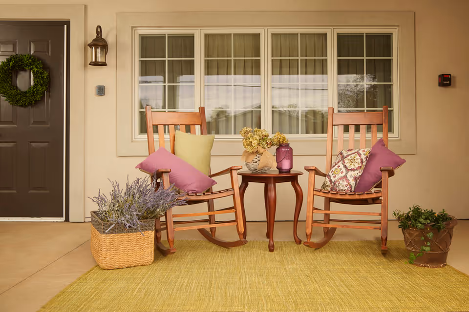 A cozy outdoor porch area with two wooden rocking chairs adorned with colorful cushions, a small round wooden table between them holding a vase with dried flowers and a purple lantern. There is a green wreath hanging on a dark brown door to the left, a basket with lavender flowers on the floor, and a potted plant on the right side. The background features a beige wall with a large window.