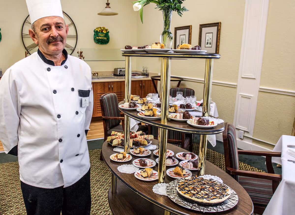 A chef in a white uniform and hat stands next to a multi-tiered serving cart filled with various plated desserts in a dining room with tables and chairs.