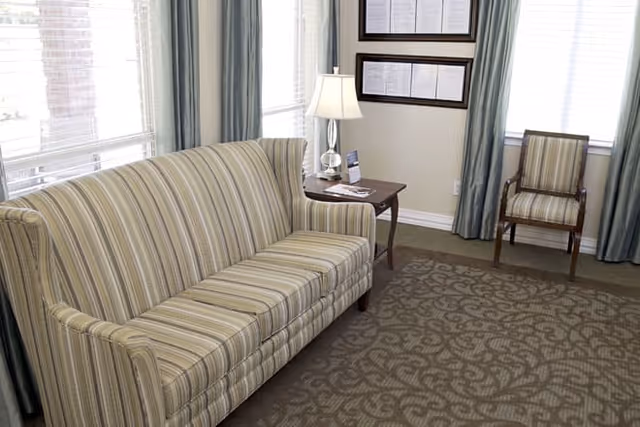 A cozy living room area with a striped sofa, a wooden side table with a lamp and some papers, a patterned carpet, and a wooden chair with striped upholstery. The room has large windows with light blue curtains allowing natural light to fill the space.