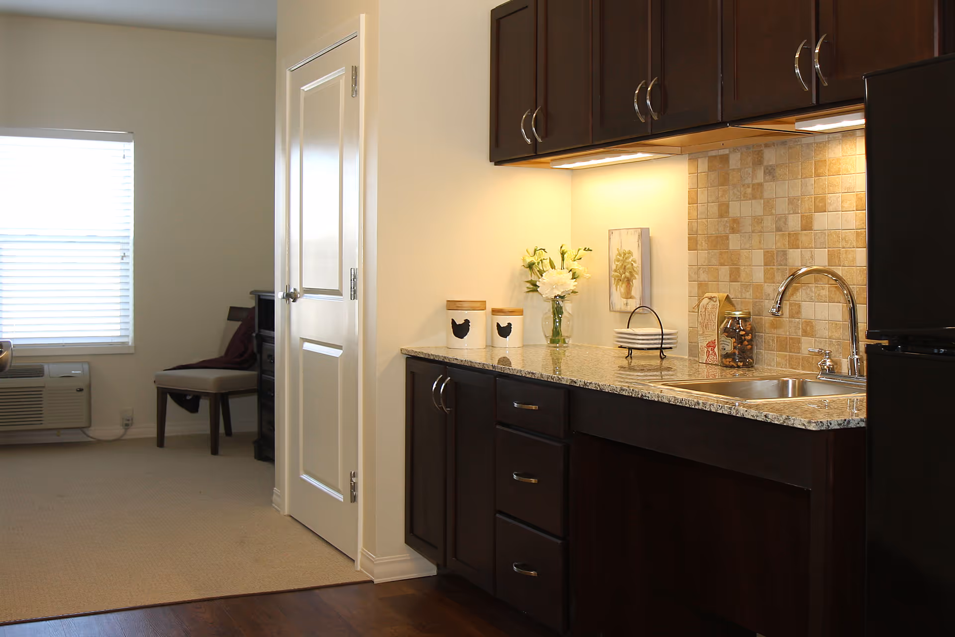 A kitchenette area with dark wood cabinets, a granite countertop, a stainless steel sink, and a tiled backsplash. On the countertop, there are two canisters with chicken silhouettes, a vase with white flowers, a small stack of plates, and a jar of candy. To the left, there is a door and a window with blinds, and a chair with a dark cushion is partially visible.