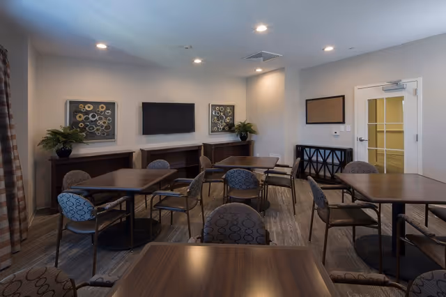 A well-lit interior room with multiple square tables and cushioned chairs arranged around them. The room features neutral-colored walls with two framed abstract artworks and a flat-screen TV mounted on the wall. There are two potted plants on dark wooden cabinets beneath the artworks. A door with glass panels is visible on the right side, and the floor is carpeted in a patterned design.
