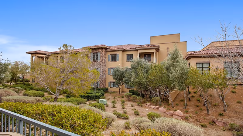 Exterior view of a multi-story senior living facility building with beige and stone facade, surrounded by landscaped bushes, trees, and rocks under a clear blue sky.