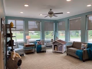 Sunlit common room with sofas and window seats, a small doghouse, a bookshelf, side tables, and a ceiling fan.