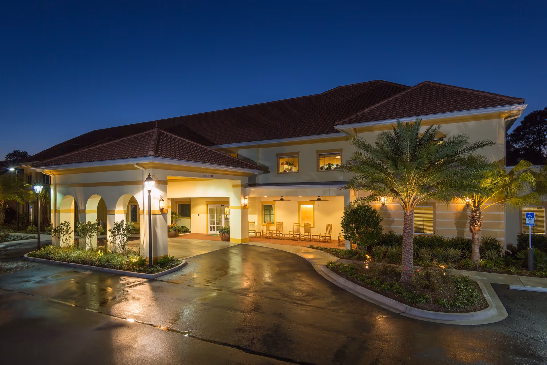 Exterior view of The Windsor Of Gainesville senior living facility at dusk, showing the entrance with a covered driveway, warm lighting, palm trees, and a landscaped area with bushes and plants.