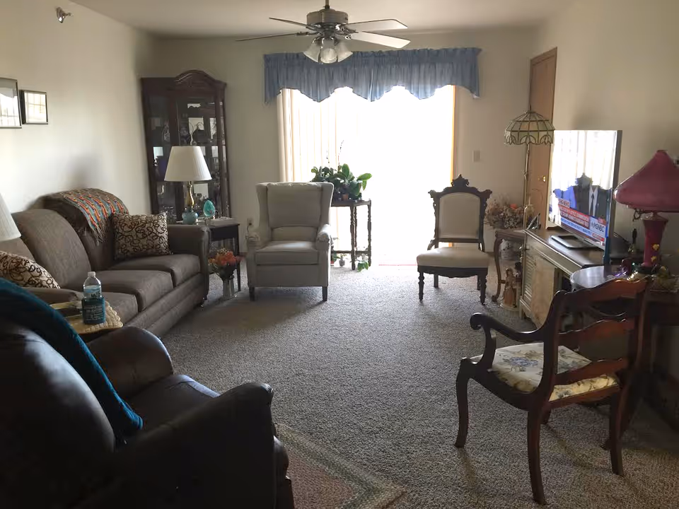 Cozy living room with sofa and multiple armchairs facing a TV, a glass-front cabinet, ceiling fan, and a sliding glass door letting in light.