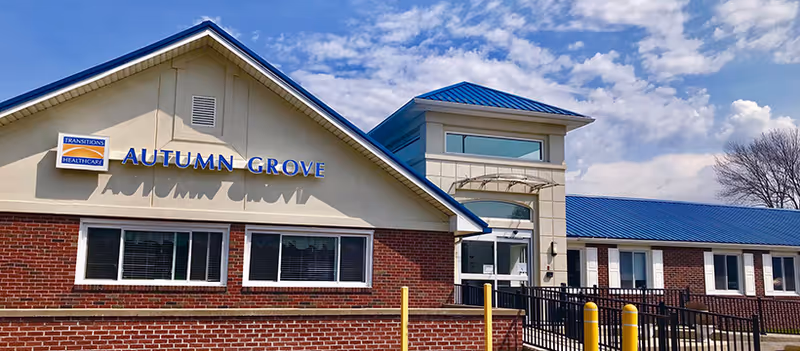 Exterior view of Transitions Healthcare Autumn Grove building with a brick facade, beige upper walls, blue metal roof, and a clear sky with some clouds.