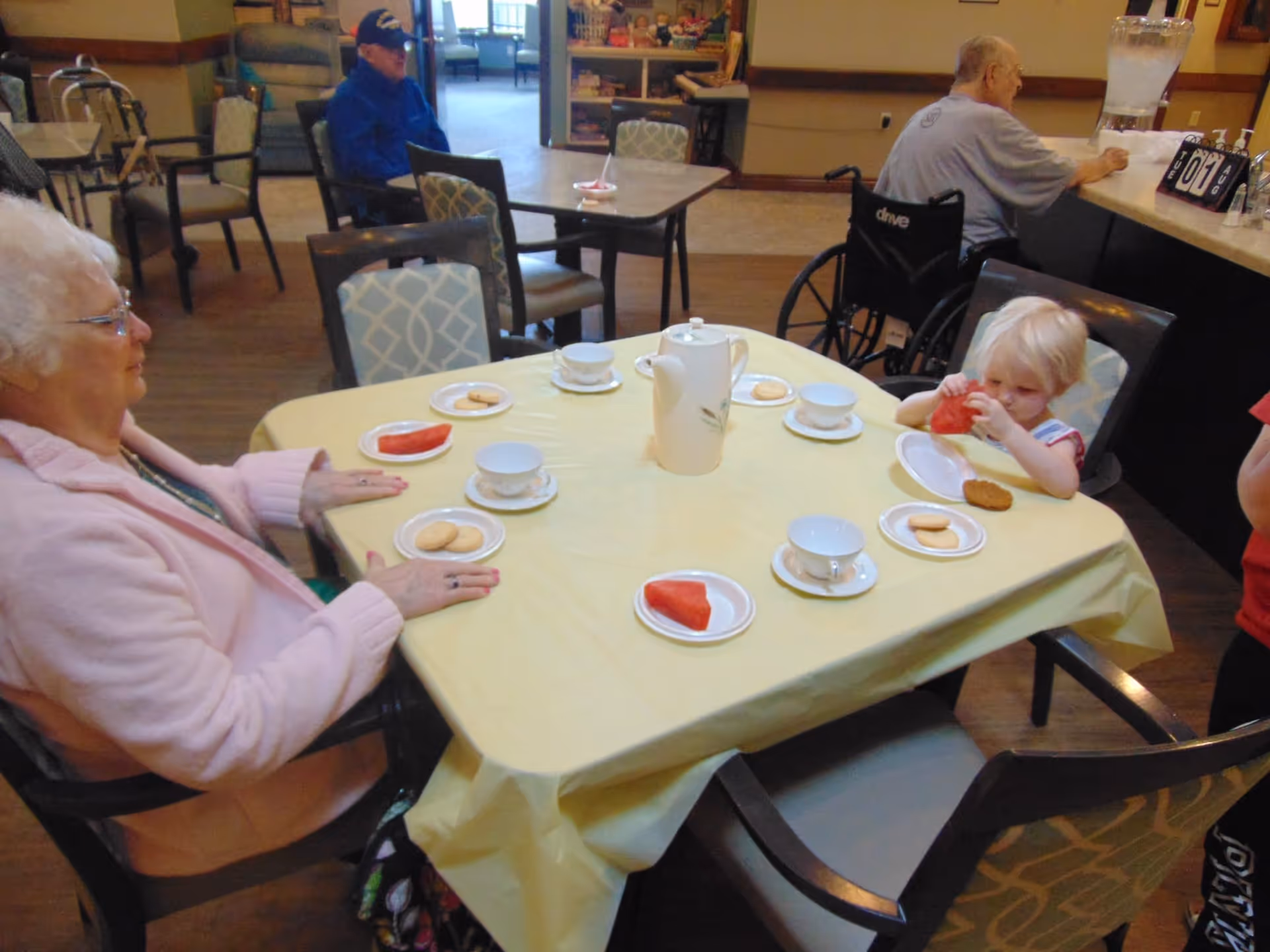 An elderly woman and a young child sit at a table covered with a yellow tablecloth in a communal dining area. The table has plates with cookies and watermelon slices, teacups, and a teapot. In the background, an elderly man in a wheelchair is seated at a counter, and another man in a blue jacket and cap is sitting at a table further back. The room has wooden flooring and several chairs with patterned upholstery.