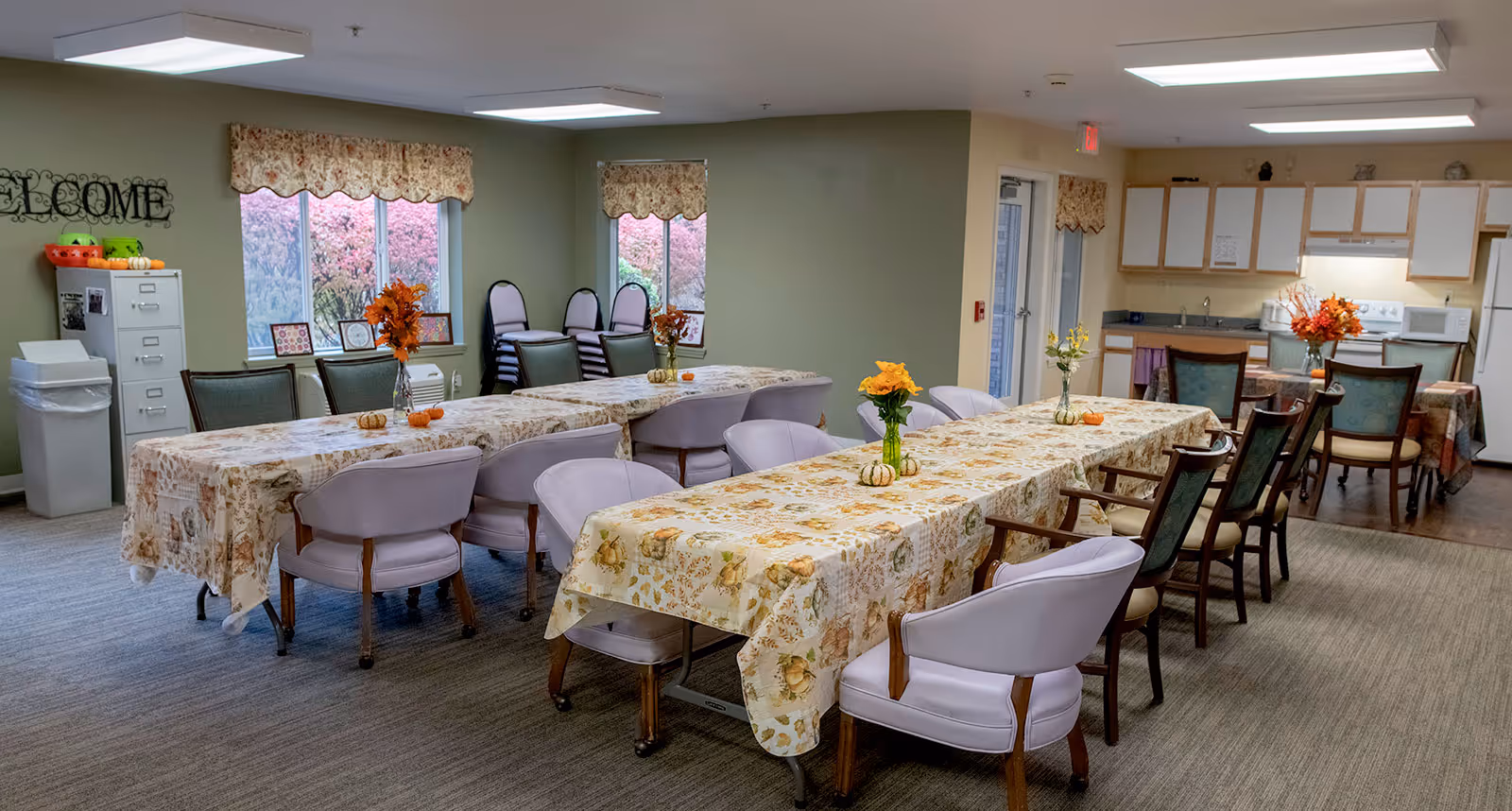 A dining area in a senior living facility with two long tables covered in floral tablecloths and decorated with small vases of flowers and miniature pumpkins. The room has multiple chairs around the tables, a small kitchen area with cabinets, a refrigerator, microwave, and stove in the background. There are windows with floral valances and a filing cabinet with a trash bin nearby. The walls are painted light green and beige.