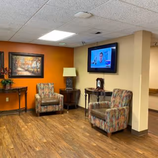 A cozy seating area in a senior living facility with two patterned armchairs, a small wooden table with a lamp, a wall-mounted flat screen TV, a framed autumn landscape painting on an orange accent wall, and wood flooring.