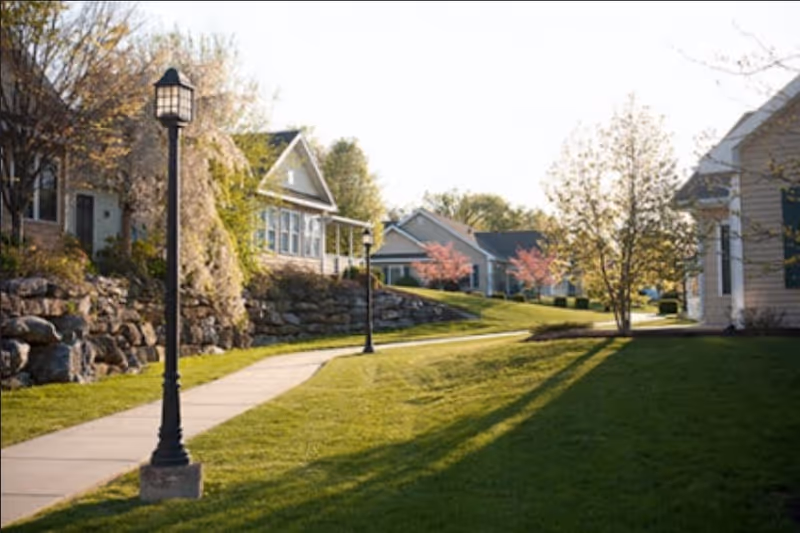 A sunny outdoor pathway lined with lamp posts and surrounded by well-maintained grass, trees, and residential-style buildings with stone retaining walls and blooming trees.