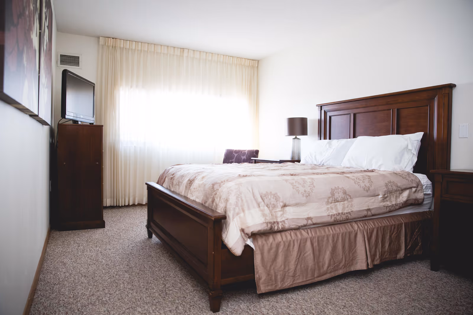 A bedroom with a large wooden bed frame, a beige patterned comforter, and white pillows. There is a wooden nightstand with a lamp on the right side of the bed, a wooden dresser with a flat-screen TV on the left side near a window covered with sheer white curtains, and a carpeted floor.