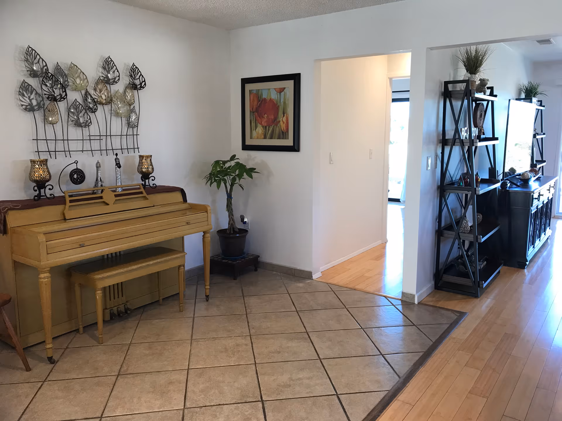 Interior view of a living area featuring a yellow upright piano with a matching bench, decorative metal leaf wall art above the piano, two candle holders, a potted plant on a small stand, a framed floral painting on the wall, and a black shelving unit with various decorative items and a TV in the background. The floor transitions from tiled to wood.