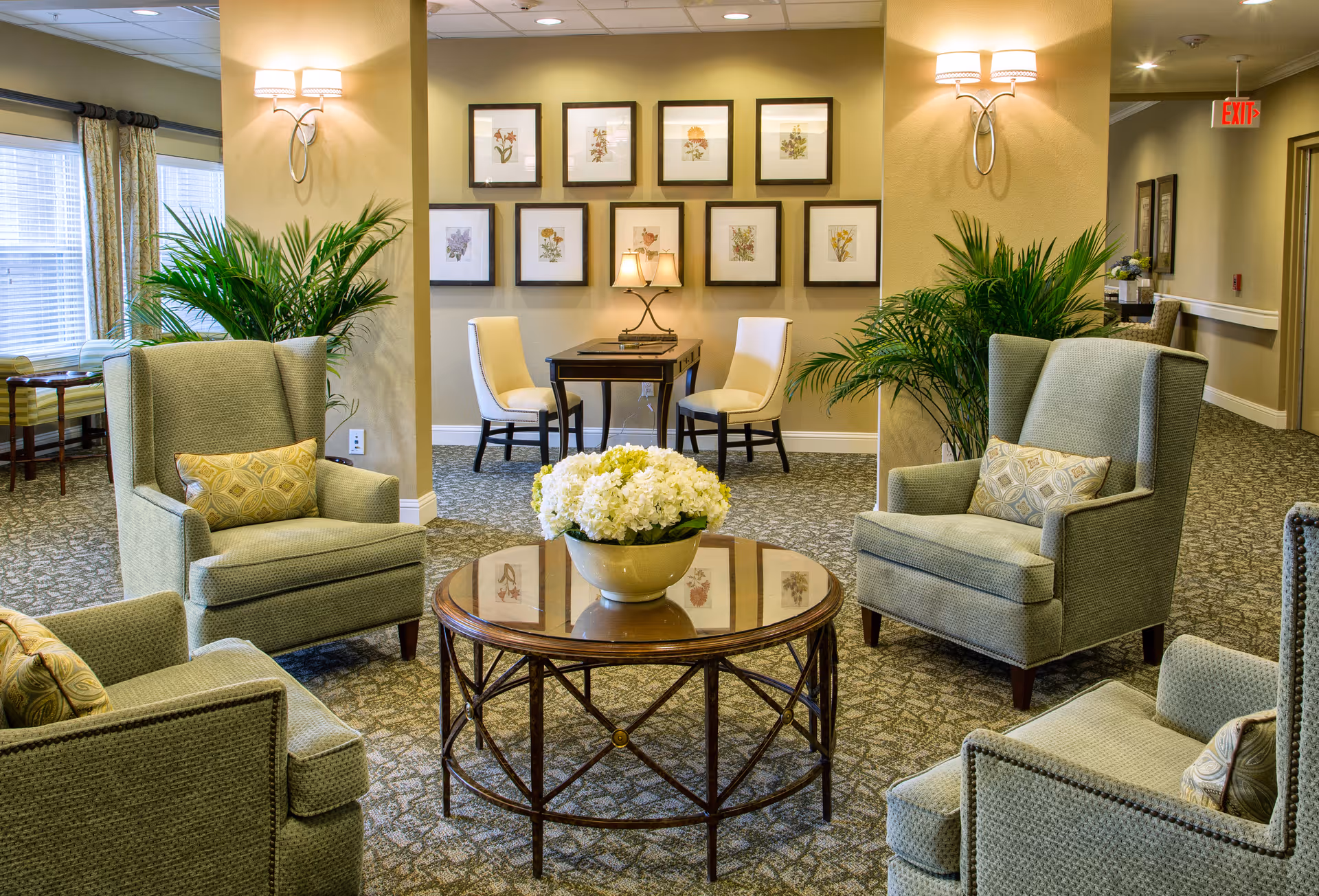 A comfortable senior living lounge with four upholstered armchairs arranged around a round glass-top coffee table holding a floral centerpiece, framed artwork and potted plants.