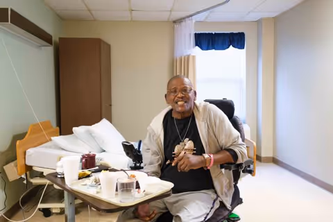 An elderly man sitting in a wheelchair in a bedroom with a hospital bed, eating a meal on a tray table. The room has beige walls, a window with curtains, and a wooden cabinet.